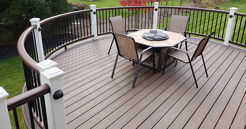 A wooden deck with a curved railing, hosting a patio dining table and chairs, overlooking a grassy lawn.